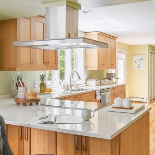 Open floor plan kitchen with large island, wood cabinets, and cooktop with overhead range hood (JM Construction)